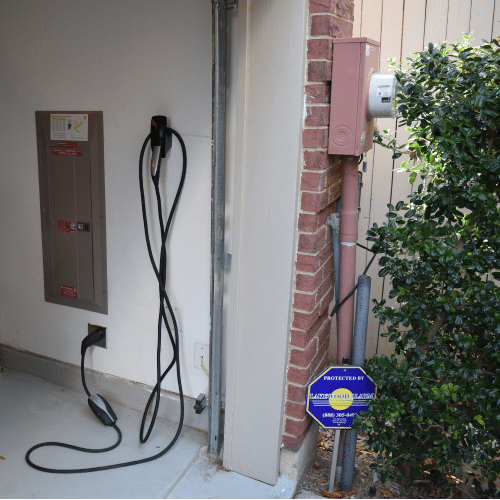 A garage corner showing an electrical panel, EV charger cable hanging on the wall, and a security sign next to a bush.